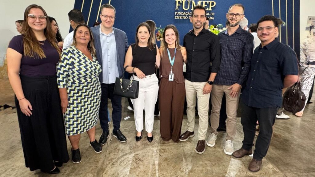 A imagem mostra um grupo de sete pessoas posando para uma foto em um ambiente interno. O grupo é composto por quatro mulheres e três homens, todos vestidos de forma casual e elegante. Eles estão alinhados lado a lado, sorrindo para a câmera. Ao fundo, há uma parede azul escura com faixas verticais douradas e o texto em dourado "FUMEP FUNDAÇÃO MUNICIPAL DE ENSINO DE PIRACICABA", indicando que o local está relacionado a uma instituição educacional municipal da cidade de Piracicaba. O chão é claro e parece ser de cimento polido ou pedra. A iluminação é clara e uniforme, destacando bem os rostos e roupas das pessoas. O ambiente transmite uma sensação de formalidade moderada, possivelmente durante um evento ou reunião oficial da fundação.