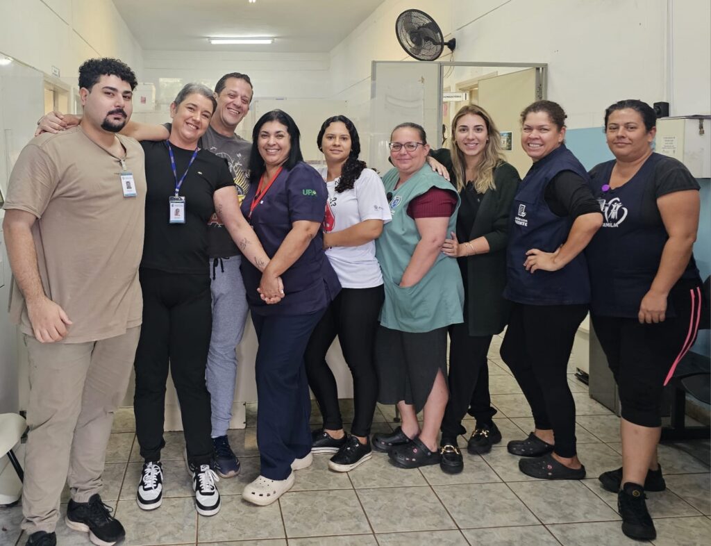 A imagem mostra um grupo de nove pessoas, compostas por homens e mulheres, posando juntas em um corredor interno que parece ser de uma unidade de saúde. O ambiente é bem iluminado com luz artificial branca e as paredes são claras, conferindo um aspecto limpo e organizado ao local. Algumas pessoas usam uniformes médicos, como jalecos e camisetas com a sigla "UPA" (Unidade de Pronto Atendimento), indicando que trabalham na área da saúde. Outras vestem roupas casuais ou coletes identificados, possivelmente de equipes administrativas ou de apoio. Ao fundo, há portas com placas, uma delas indicando "CONSULTÓRIO", reforçando o contexto clínico do local. Também é possível ver um ventilador de parede. O grupo está alinhado e sorridente, transmitindo um clima de amizade e colaboração profissional. A imagem sugere uma equipe unida e dedicada, provavelmente pronta para atender ou já em atividade na unidade de saúde.