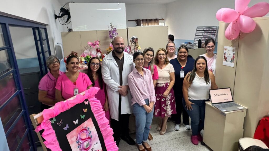 A imagem mostra um grupo de onze pessoas, sendo dez mulheres e um homem, posando para uma foto em um ambiente interno que parece ser um escritório ou uma clínica. O homem está vestido com um jaleco branco, sugerindo que ele pode ser um profissional da área da saúde. As mulheres estão vestidas em tons claros, com várias usando rosa, o que pode estar relacionado à decoração temática do local. Ao lado esquerdo do grupo, há uma grande placa decorativa com a forma do número 8, adornada com flores e uma borda de papel rosa, indicando que a celebração é provavelmente em homenagem ao Dia Internacional da Mulher. No fundo, há uma parede com divisórias, um ventilador de parede, um relógio digital e uma mesa com um laptop que exibe a frase "Café das Mulheres", reforçando o tema da comemoração. Também é possível ver balões rosa em formato de flor presos a uma das divisórias. O ambiente transmite uma atmosfera alegre e acolhedora, com as pessoas sorrindo e aparentando estar felizes em participar do evento. Há ainda cestas ou arranjos florais sobre um balcão atrás do grupo, complementando a decoração festiva.