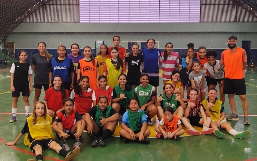 A imagem mostra um grupo grande de meninas jovens, cerca de 25, reunidas em uma quadra esportiva coberta, provavelmente em um ginásio. Elas estão vestindo roupas esportivas, muitas com coletes coloridos nas cores amarelo, laranja, verde e azul, todos com a inscrição "SELAM". Algumas meninas estão sentadas no chão na frente, outras estão agachadas no meio, e o restante está em pé atrás, formando várias fileiras para a foto em grupo. Ao lado direito da foto, há um adulto, provavelmente o treinador, vestido com regata laranja e shorts pretos. A quadra tem piso verde com marcações brancas e vermelhas, e o ambiente é iluminado por luz artificial, com paredes cinza e teto alto com estrutura metálica. O clima da foto é descontraído e alegre, com as meninas sorrindo e algumas fazendo gestos com as mãos, demonstrando entusiasmo e união do grupo. A palavra "SELAM" sugere que o grupo faz parte de um programa ou equipe esportiva ligada à Secretaria de Esportes.