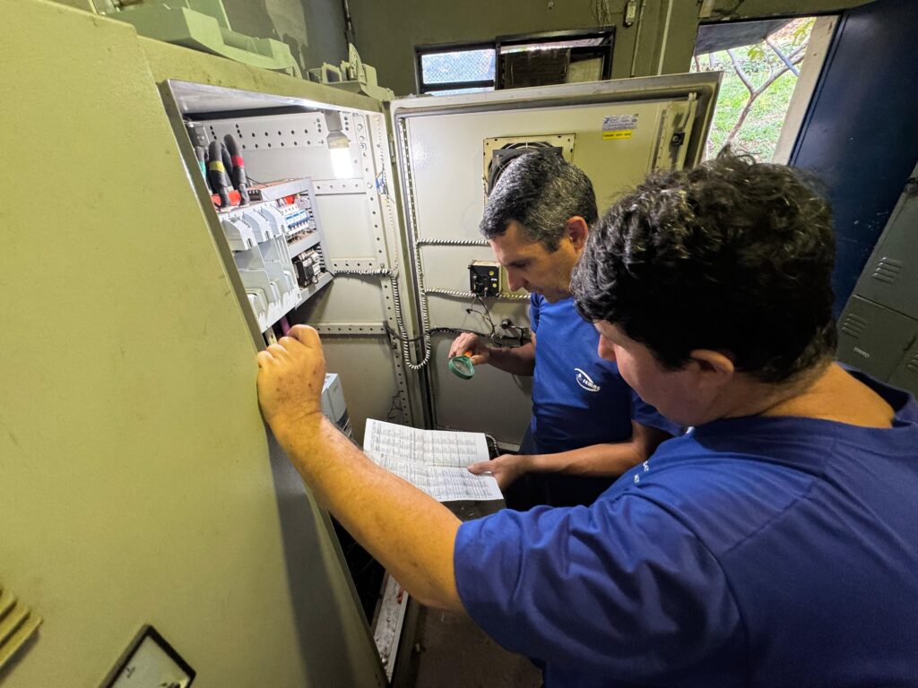 A imagem mostra dois técnicos, ambos vestidos com camisas azuis com o logo "SEMAE", trabalhando em um painel elétrico aberto. Eles estão em uma sala industrial, com uma janela ao fundo que revela uma área externa com vegetação. O painel elétrico é metálico e contém diversos componentes internos, como disjuntores, fios e um ventilador de resfriamento. Um dos técnicos está segurando um documento impresso, provavelmente um manual ou esquema técnico, enquanto o outro usa uma ferramenta verde, talvez uma lupa ou sensor, para inspecionar os componentes do painel. Há uma etiqueta amarela no painel que indica "TENSÃO - 220V / 60HZ". A cena transmite um ambiente de manutenção ou inspeção técnica em infraestrutura elétrica, com foco na precisão e segurança do trabalho.