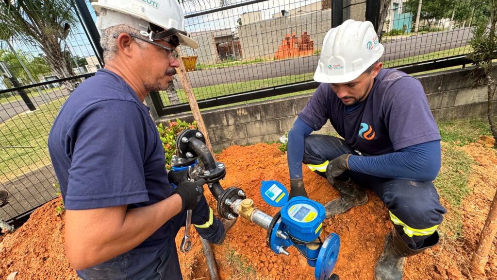 A imagem mostra dois trabalhadores homens instalando ou realizando manutenção em um sistema de infraestrutura de água. À esquerda, um homem com cabelo grisalho, usando óculos e capacete branco, está em pé e utilizando uma chave inglesa em uma tubulação metálica preta. Ele veste uma camiseta azul escura e luvas pretas. À direita, um homem mais jovem, com barba, também usando capacete branco e camiseta azul escura com um logotipo circular, está agachado observando um hidrômetro digital azul com a tampa aberta. Ambos usam calças escuras com faixas refletivas amarelas e botas escuras. O trabalho ocorre em um terreno de terra vermelha. Ao fundo, há uma cerca metálica preta, vegetação, uma via pavimentada e um prédio em construção com tijolos aparentes. A iluminação é natural e intensa, indicando que a foto foi tirada durante o dia. O destaque visual fica por conta do hidrômetro azul contrastando com a terra vermelha.
