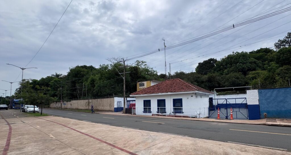 A imagem mostra uma pequena construção branca de um andar com janelas e base pintadas de azul escuro, além de um telhado de telhas vermelhas. Na frente do prédio há um corrimão branco e uma calçada. Ao lado direito do prédio há um portão azul com duas cones laranja em frente. A construção está localizada ao lado de uma rua asfaltada com uma linha amarela no centro. Do lado oposto da rua, há uma calçada com uma faixa pintada de vermelho. No fundo, vê-se uma área verde com muitas árvores e um poste de energia com vários fios. O céu está nublado e o clima parece ser tranquilo. Também é possível ver um carro branco e um caminhão azul estacionados, e uma pessoa caminhando na calçada ao longe.