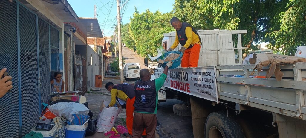 A imagem mostra uma ação comunitária de limpeza em uma rua residencial, provavelmente no município de Piracicaba, Brasil, voltada para o combate à dengue, conforme indicado pela faixa na lateral do caminhão que diz "ARRASTÃO DENGUE 2022". Três trabalhadores municipais, vestidos com uniformes amarelos e laranjas, estão recolhendo lixo e objetos que podem acumular água parada, como caixas plásticas e sacos, para evitar a proliferação do mosquito Aedes aegypti, transmissor da dengue. Um dos trabalhadores está dentro da carroceria do caminhão, recebendo os materiais recolhidos por outro homem, enquanto um terceiro está agachado, organizando o que foi coletado. Ao fundo, uma menina observa a ação da porta de uma casa. A rua é estreita e ladeada por casas simples, com árvores e um céu azul claro ao fundo. A cena transmite uma sensação de trabalho comunitário e esforço conjunto para a saúde pública.