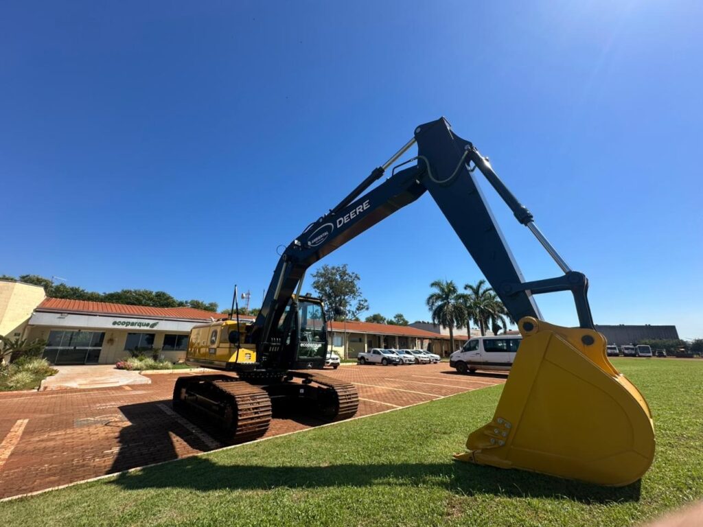 A imagem mostra uma escavadeira grande da marca John Deere, com cores predominantes amarelo e preto, estacionada em um local aberto que combina piso pavimentado e grama verde. A escavadeira está posicionada de forma que seu braço está levantado e curvado para baixo, com a pá tocando a grama. A foto foi tirada de um ângulo baixo, o que dá uma sensação de imponência e grande porte à máquina. Ao fundo, há um prédio com uma placa escrita "ecoparque" e um telhado com telhas vermelhas. Também é possível ver alguns veículos brancos estacionados e palmeiras, indicando um ambiente possivelmente tropical ou subtropical. O céu está claro e ensolarado, com luz forte que projeta sombras da escavadeira no chão, sugerindo que a foto foi tirada durante o dia. A escavadeira tem adesivos e logotipos, incluindo "DEERE", "AMBIENTAL" e "ecoparque", indicando que provavelmente é utilizada para serviços ambientais ou de manutenção em áreas verdes. A imagem transmite uma sensação de robustez e eficiência da máquina em um ambiente de trabalho ao ar livre.