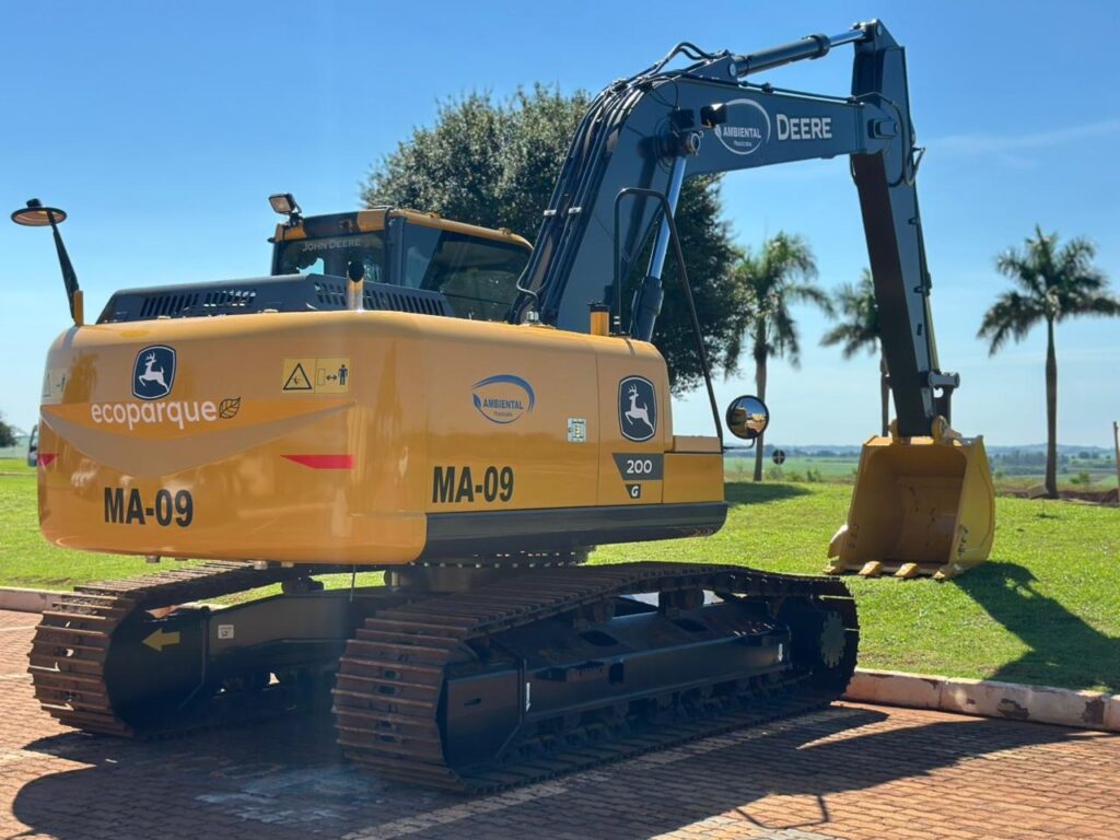 A imagem mostra uma escavadeira hidráulica da marca John Deere, modelo 200G, em destaque. A máquina é predominantemente amarela com detalhes em preto, e está posicionada em um terreno com grama e próximo a um pavimento de blocos intertravados de cor avermelhada. No braço da escavadeira, há inscrições como "ecoparque" e "AMBIENTAL Piracicaba", indicando que provavelmente pertence a uma empresa ou órgão ambiental da cidade de Piracicaba. Ao fundo, vê-se um céu azul claro e algumas palmeiras, sugerindo que a foto foi tirada em um dia ensolarado. A escavadeira está parada, com a lança levantada e o balde apoiado no chão, transmitindo uma sensação de robustez e funcionalidade.