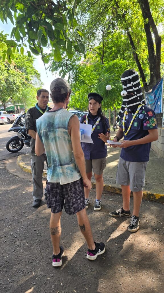 A imagem mostra quatro pessoas em um espaço ao ar livre, provavelmente em um parque ou próximo a uma rua, durante o dia. Três delas estão vestidas com uniformes que lembram escoteiros, com camisas azul-escuras, lenços amarelos e brancos no pescoço e bermudas cinza. Uma dessas pessoas está usando uma máscara alta, listrada em preto e branco, com grandes olhos brancos, que chama bastante atenção. Essa pessoa segura alguns papéis e parece estar conversando com um homem que está de costas para a câmera. Esse homem veste uma regata tie-dye colorida, bermuda com estampa camuflada e um boné também camuflado. À esquerda, há outro homem com uniforme verde-escuro e calça cinza, que observa a interação. Ao fundo, é possível ver árvores, um motocicleta e alguns carros estacionados. A luz do dia ilumina a cena, criando um ambiente descontraído e informal.