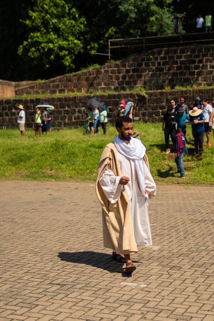 A imagem mostra um homem vestido com roupas que lembram trajes bíblicos: túnica branca longa, manto bege e um xale branco enrolado no pescoço. Ele está caminhando sobre um piso de pedras retangulares cinzas. Ao fundo, há um muro de pedras e uma grade metálica, além de um grupo de pessoas vestindo roupas modernas, como camisetas e chapéus, algumas segurando guarda-sóis. O cenário parece ser um local histórico ou arqueológico ao ar livre, com áreas de grama verde e árvores ao fundo. A iluminação é forte, indicando um dia ensolarado, e a foto está focada no homem, com o fundo ligeiramente desfocado. A cena sugere uma encenação ou representação histórica, possivelmente bíblica.