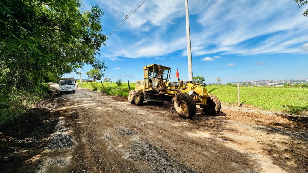 A imagem mostra uma cena de manutenção de estrada em uma área rural durante o dia. No centro da foto, há uma motoniveladora amarela grande, marca Caterpillar, modelo 120K, que está trabalhando sobre uma estrada de terra. Na parte traseira da motoniveladora, há um cone de sinalização laranja colocado sobre a máquina. À esquerda da imagem, mais ao fundo, um caminhão branco da marca Mercedes-Benz está parado ou se aproximando da motoniveladora. Ao redor, há vegetação verde densa, com árvores altas à esquerda e um campo gramado com cerca ao lado direito da estrada. O céu está azul e limpo, indicando um dia ensolarado. A cena sugere que está sendo realizado um trabalho de nivelamento ou manutenção da estrada de terra, em um ambiente rural e provavelmente em clima tropical ou subtropical, devido à vegetação presente.