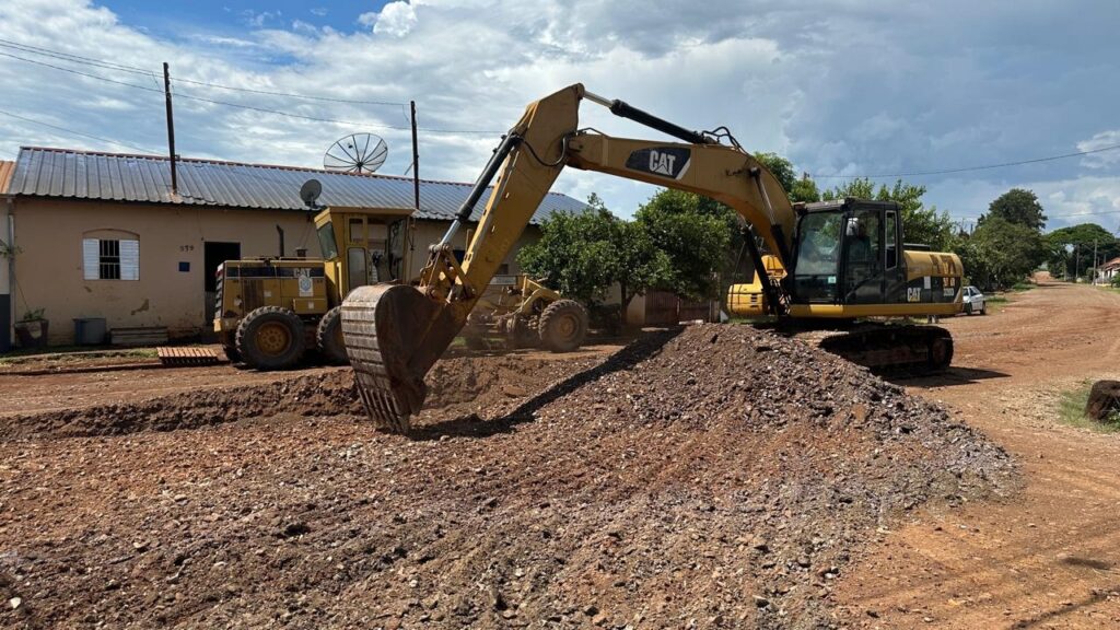 A imagem mostra uma cena rural durante o dia, com o céu parcialmente nublado. No centro da imagem, há uma escavadeira amarela da marca Caterpillar (CAT) posicionada sobre uma pilha de terra escura e pedregosa, cavando uma vala que atravessa uma estrada de terra. À esquerda, um nivelador de estrada amarelo, também da marca CAT, está estacionado. Ao fundo, há uma casa simples de um andar, de cor bege, com telhado de metal ondulado, uma antena parabólica e uma pequena janela. Árvores verdes e postes de energia elétrica alinham-se ao longo da estrada, que se estende para o lado direito da imagem. A iluminação é natural e clara, com sombras suaves. A cena transmite uma sensação de trabalho em andamento em um ambiente rural, com máquinas pesadas em ação para construção ou manutenção da estrada.
