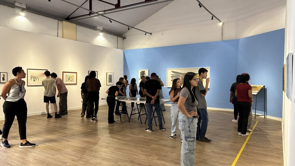 A imagem mostra uma galeria de arte com um grupo de pessoas, principalmente jovens, explorando o espaço. O ambiente é amplo, com paredes brancas e azul claro, teto alto com vigas metálicas expostas e piso de madeira clara. As pessoas estão distribuídas pelo local, algumas observando quadros nas paredes e outras em volta de uma mesa central com uma superfície de vidro, possivelmente exibindo objetos ou documentos. A iluminação é clara e bem distribuída, vinda de luminárias no teto. No lado direito do espaço, há uma linha amarela no chão que provavelmente delimita a área para os visitantes. A atmosfera parece tranquila e contemplativa, típica de um museu ou galeria de arte.