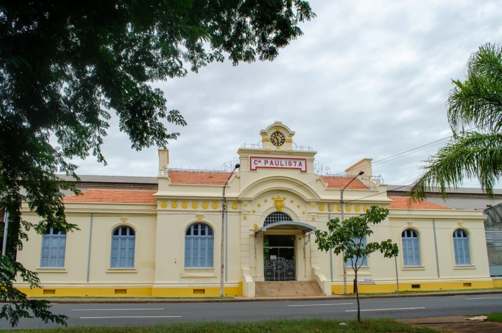 A imagem mostra a fachada de um prédio antigo de estilo colonial, provavelmente uma antiga estação ferroviária. O edifício é pintado em um tom claro, com detalhes em amarelo vibrante na base e ao redor das janelas. No centro da construção, há uma porta de entrada com um arco e um pequeno toldo metálico, acima da qual está uma placa com o texto "Cia PAULISTA" em letras vermelhas e um relógio analógico circular. O prédio possui janelas arqueadas com persianas azuis e um telhado de telhas vermelhas. Em frente ao edifício, há uma rua asfaltada com sinalização branca. A cena é enquadrada por galhos verdes de árvores, e o céu está nublado, conferindo uma iluminação suave à fotografia. O conjunto transmite uma atmosfera histórica e preservada, típica de construções antigas ligadas à ferrovia no Brasil.