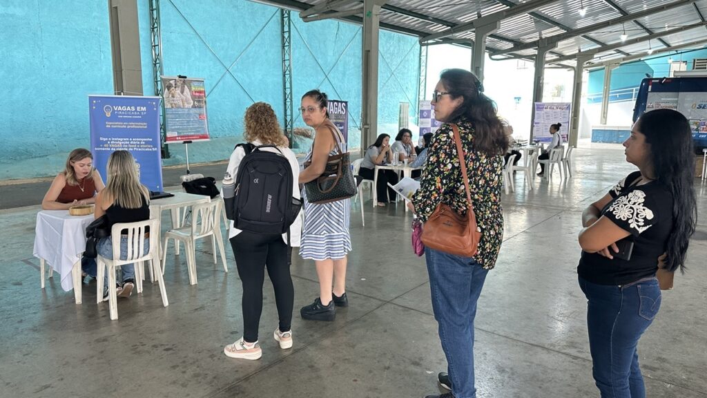 A imagem mostra um evento de empregabilidade ou uma feira de empregos realizada em um espaço amplo e coberto, com estrutura metálica e paredes azuis claras. No centro, várias mulheres estão em fila ou interagindo em mesas de atendimento. À esquerda, uma mulher loira vestindo uma blusa vermelha está sentada atrás de uma mesa branca, conversando com outra mulher que está de costas para a câmera. Ao lado, na fila, há várias mulheres, uma delas com mochila preta, outra de vestido listrado azul e branco, e outras vestidas de forma casual. No fundo, há outras mesas com pessoas sentadas e atendendo o público. Na parede, há um banner azul com o texto "VAGAS EM PIRACICABA SP COM ADRIANA CASARIN", que indica que o evento está focado em oportunidades de emprego na cidade de Piracicaba, São Paulo. O banner também destaca que Adriana Casarin é especialista em reformulação de currículos e que divulga diariamente mais de 40 vagas na cidade. O ambiente é iluminado naturalmente, transmitindo uma atmosfera de organização e profissionalismo.