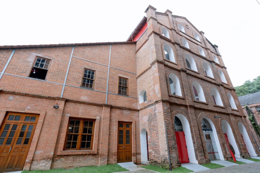 A imagem mostra um prédio grande e antigo, construído em tijolos vermelhos. O edifício tem duas partes principais: uma ala mais alta, com cinco andares, e outra mais baixa, com dois andares. A ala alta possui várias janelas arqueadas, pintadas de branco no interior, e grandes portas arqueadas no térreo, uma delas vermelha. A ala mais baixa tem janelas retangulares com molduras de madeira e uma porta dupla de madeira. O telhado é inclinado e revestido com telhas. O céu está nublado, o que deixa a iluminação suave e uniforme. Na frente do prédio, há uma faixa de grama e um caminho pavimentado. A foto foi tirada de um ângulo baixo, destacando a altura do edifício.