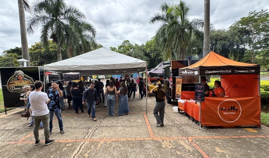 A imagem mostra uma feira de alimentos ao ar livre, com várias barracas e food trucks dispostos em um espaço pavimentado com detalhes em tijolos vermelhos. O ambiente é arborizado, com árvores verdes e algumas palmeiras ao fundo, sugerindo um local agradável e com sombra. O céu está nublado, o que proporciona uma iluminação suave e difusa. No centro da imagem, várias pessoas estão caminhando ou paradas, observando as barracas. Algumas estão conversando, enquanto outras aguardam atendimento. À esquerda, há um food truck preto com o nome "Dona S." e uma tenda branca ao lado. À direita, destaca-se uma barraca laranja da "Picolle Sorvetes", que oferece sorvetes, açaí e milk shakes, conforme indicado nas placas. As pessoas estão vestidas de forma casual, com roupas leves, como camisetas, calças e saias, transmitindo uma atmosfera descontraída e amigável, típica de eventos comunitários ou festivais de rua. A composição da foto mostra uma movimentação ativa, com um clima de confraternização e lazer.