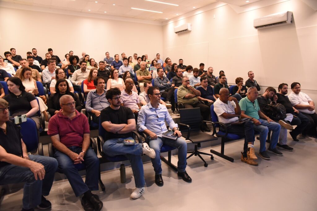 A imagem mostra um grande grupo de pessoas sentadas em um auditório ou sala de aula com assentos em níveis escalonados. São cerca de 50 a 60 pessoas, predominantemente homens de meia-idade, vestindo roupas casuais ou semi-formais, como camisetas, camisas pólo e camisas sociais. A maioria parece estar atenta, possivelmente assistindo a uma palestra ou apresentação. O ambiente é bem iluminado, com paredes brancas e cadeiras azuis com braços pretos. No canto direito superior da sala, há dois aparelhos de ar condicionado instalados na parede. O clima geral da imagem transmite um ambiente profissional, como um seminário, workshop ou aula acadêmica.