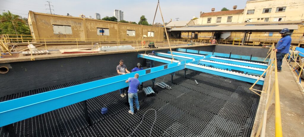 A imagem mostra um ambiente industrial, provavelmente uma estação de tratamento de água, onde um grupo de trabalhadores está realizando uma instalação ou manutenção dentro de um tanque retangular de concreto. No interior do tanque, há uma estrutura em forma de grade preta, que parece ser um sistema de filtragem ou suporte. Sobre essa estrutura, estão sendo posicionadas vigas metálicas azuis, que estão sendo suspensas por um guindaste, visível através do cabo pendurado. Cinco trabalhadores estão visíveis na cena: quatro dentro do tanque, envolvidos diretamente na instalação das vigas, e um do lado de fora, em cima da borda do tanque, provavelmente supervisionando ou auxiliando. Os trabalhadores usam roupas comuns de trabalho, como camisetas e calças, e alguns usam capacetes de segurança. Ao fundo, é possível ver construções industriais antigas com paredes amareladas e, mais ao longe, prédios mais modernos de vários andares. O céu está claro, indicando que a foto foi tirada durante o dia. A imagem é capturada de um ponto elevado, proporcionando uma visão ampla do tanque e do trabalho em andamento. Também há guarda-corpos amarelos ao redor do tanque para segurança.