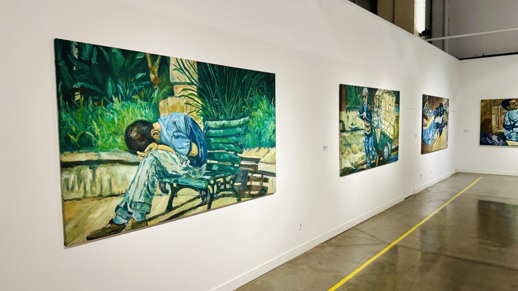 A imagem mostra o interior de uma galeria de arte com várias pinturas grandes expostas em uma parede branca. A pintura principal, à esquerda, retrata um homem sentado em um banco de parque verde, com a cabeça abaixada e apoiada nos braços, transmitindo uma sensação de cansaço ou tristeza. Ele veste uma camisa azul e calças claras. O fundo da pintura é composto por folhagens verdes e grama alta, dando uma sensação de natureza ao ambiente retratado. À direita dessa obra, há outras três pinturas visíveis, cada uma com estilos e temas variados, mas todas com cores vivas e pinceladas expressivas. O chão da galeria é de concreto polido, e há uma linha amarela no chão paralela à parede, provavelmente para orientar os visitantes. A iluminação é clara e uniforme, destacando as obras de arte expostas. A perspectiva da foto é em ângulo, mostrando a extensão da parede e as obras alinhadas ao longo dela.