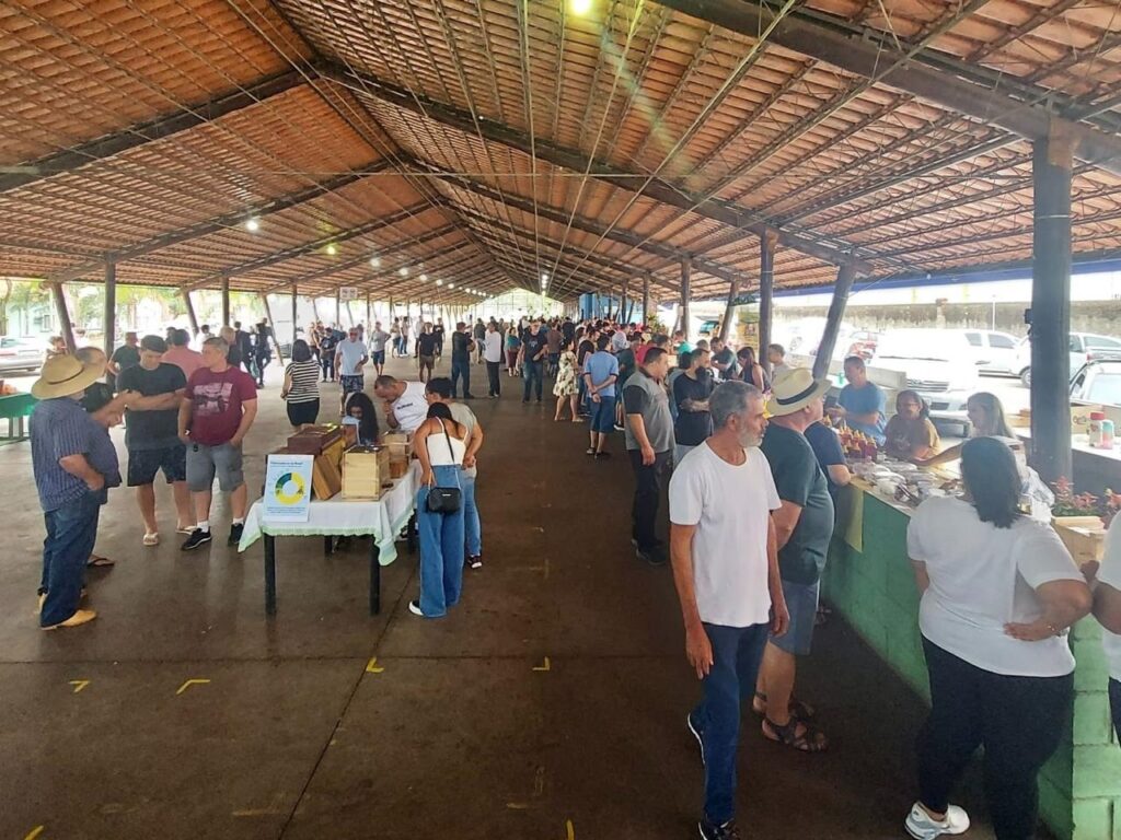 A imagem mostra um mercado ou feira coberta, bastante movimentada, localizada em um pavilhão grande com estrutura de madeira e telhado de telhas vermelhas. O ambiente é iluminado por luz natural que entra pelas laterais abertas e por pequenas lâmpadas penduradas no teto. Há várias bancas e mesas ao longo dos lados, onde pessoas estão vendendo ou exibindo produtos. Muitas pessoas circulam pelo espaço, algumas olhando as bancas, outras esperando em filas. O piso é escuro, possivelmente de concreto ou terra batida, e ao fundo é possível ver carros estacionados ao ar livre sob um céu claro. O clima é de um evento comunitário, descontraído, com pessoas vestindo roupas casuais, como camisetas, shorts e chapéus. O local parece ser um ponto de encontro para compras locais, possivelmente uma feira de agricultores ou artesanato.