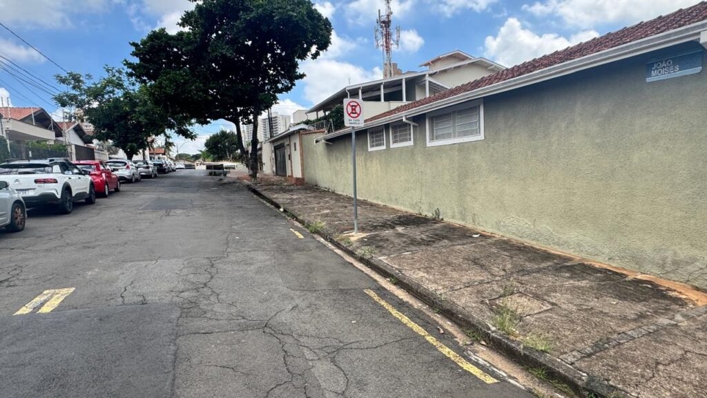 A imagem mostra uma rua residencial tranquila, provavelmente em uma cidade brasileira, dado o nome da rua "João Moisés" e o texto em português. À direita, há um longo muro pintado de verde claro que acompanha a calçada, que aparenta estar um pouco desgastada, com algumas plantas crescendo nas frestas. À esquerda, há uma fila de carros estacionados, incluindo um Citroën C4 Cactus branco em destaque. No fundo, pode-se ver uma torre de telecomunicações que se destaca contra o céu azul com algumas nuvens brancas dispersas. Há uma placa azul indicando o nome da rua "JOÃO MOISÉS" e uma placa menor com um símbolo de "proibido estacionar" e a frase "NA FAIXA AMARELA", sugerindo que o estacionamento é proibido na faixa amarela da via. A iluminação é natural e intensa, indicando que a foto foi tirada durante o dia, provavelmente próximo ao meio-dia. A rua é asfaltada e parece ser calma, sem presença de pedestres ou veículos em movimento no momento da foto. A perspectiva da imagem cria uma linha de fuga que leva o olhar para o horizonte, dando profundidade à cena.