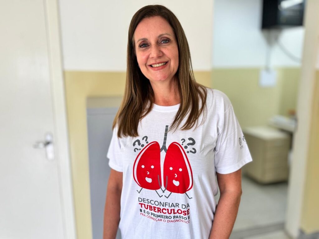 A imagem mostra uma mulher sorridente, de meia-idade, com cabelos castanhos longos, vestindo uma camiseta branca. Na camiseta, há uma ilustração de pulmões vermelhos com rostos sorridentes, acompanhada de um texto em português que diz: "DESCONFIAR DA TUBERCULOSE É O PRIMEIRO PASSO PRA COMEÇAR O DIAGNÓSTICO". Ela está em um ambiente interno, possivelmente uma clínica ou consultório, com iluminação suave e fundo desfocado, o que destaca a mulher em primeiro plano. A expressão dela é amigável e transmite uma mensagem positiva relacionada à conscientização sobre a tuberculose.