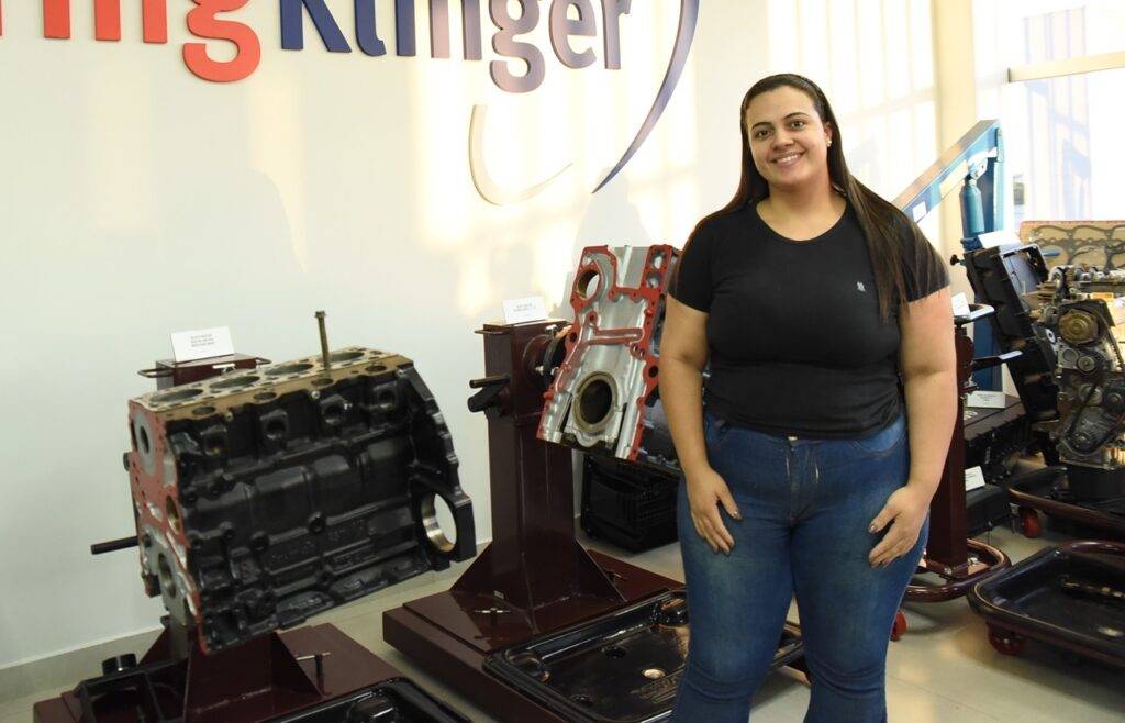 A imagem mostra uma mulher sorridente, com cabelo longo e escuro, vestindo uma camiseta preta e jeans azul, posando em um ambiente interno que parece ser uma sala de exposição ou centro de treinamento técnico. Ao fundo, há vários blocos de motor e componentes automotivos expostos sobre suportes vermelhos. Também é possível ver uma estrutura azul que provavelmente é um guincho para motores. Na parede ao fundo, está parcialmente visível o logotipo da empresa "ElringKlinger", sugerindo que o local pertence ou está associado a essa marca, que atua no setor automotivo. O ambiente é bem iluminado e organizado, com rótulos brancos próximos às peças, indicando uma apresentação detalhada dos componentes. A composição da imagem transmite um ambiente profissional e técnico voltado para motores e peças automotivas.