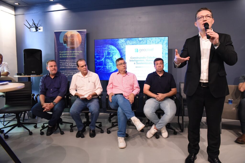 A imagem mostra uma apresentação formal em um ambiente interno, possivelmente um auditório ou sala de conferências. Um homem está em pé, à direita, falando ao microfone, usando um terno preto e camisa branca, demonstrando entusiasmo com gestos expressivos. À sua esquerda, quatro homens estão sentados em cadeiras, prestando atenção. Eles vestem roupas casuais, como camisas e jeans. Ao fundo, há uma parede azul escura com um grande monitor que exibe um globo digitalizado e o logotipo "geopixel", acompanhado do slogan "Construindo Cidades Inteligentes, Humanas e Sustentáveis". Ao lado esquerdo da imagem, há um banner vertical com texto em português que explica a evolução do processo de aprovação de projetos e emissão de alvarás em Piracicaba, destacando a integração de um sistema especializado com geoprocessamento para maior precisão e inteligência na análise dos processos. A atmosfera é profissional e tecnológica, indicando o anúncio de uma inovação relacionada à gestão urbana e uso de tecnologia para tornar a cidade mais inteligente e sustentável.
