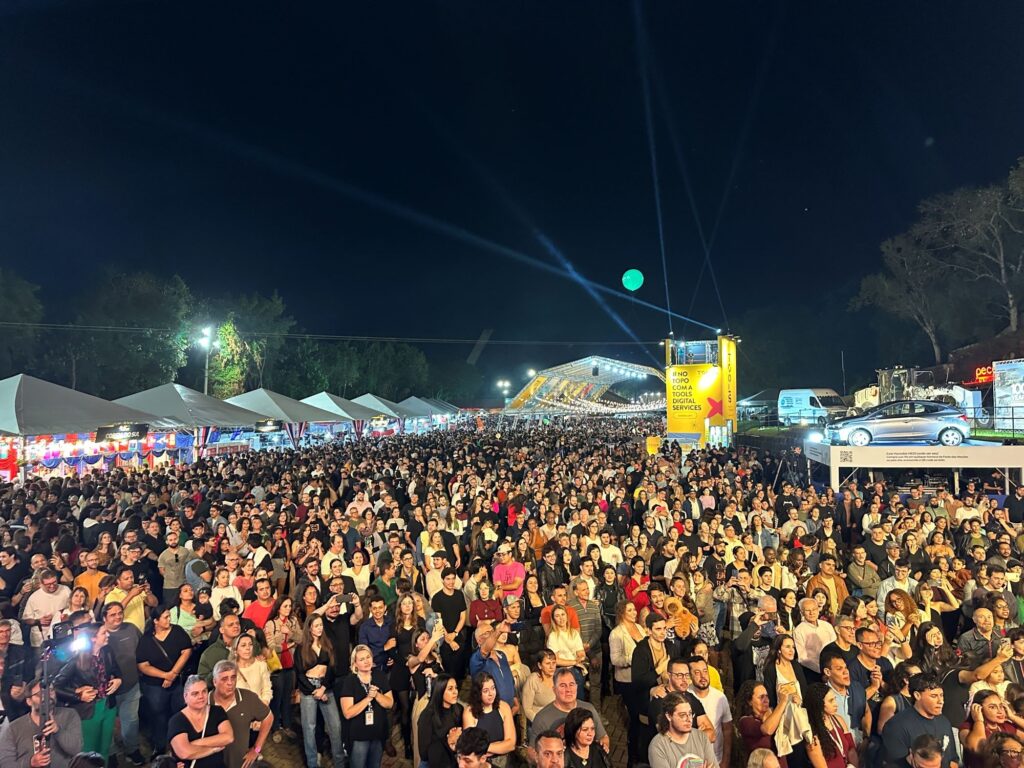 A imagem mostra uma grande festa ao ar livre à noite, com uma multidão enorme de pessoas reunidas em frente a um palco (fora do quadro, à esquerda). A multidão está bastante compacta, com pessoas vestindo roupas casuais, muitas segurando celulares para registrar o momento. À esquerda da imagem, há várias barracas brancas com toldos, provavelmente vendendo comidas, bebidas ou produtos típicos, identificadas por bandeiras coloridas. No centro, destaca-se uma torre amarela grande com o texto "#NO TOPO COM A TOOLS DIGITAL SERVICES" e a marca Hyundai, indicando que há uma rifa para ganhar um carro HB20, com instruções para compra em barracas ou via QR code. À direita, há um carro prata exposto em uma plataforma. Ao fundo, árvores e o céu escuro compõem o cenário, com luzes e holofotes iluminando a festa, criando um ambiente animado e festivo. A festa parece ser um evento cultural ou gastronômico, possivelmente chamado "Festa das Nações", onde diferentes países (como Coreia do Sul e Estados Unidos) estão representados.