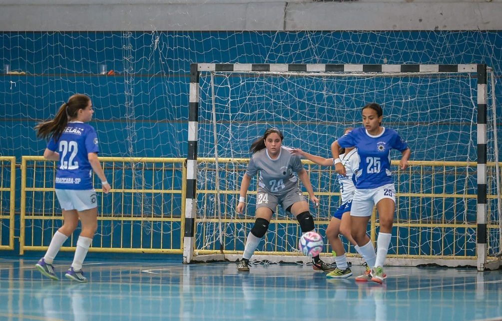 A imagem mostra uma partida de futsal feminino em ambiente interno. No centro da cena, há um gol com a rede branca e preta, e uma goleira vestindo uniforme cinza com o número 24 está em posição de defesa, com os braços abertos, focada na bola que está próxima ao chão à sua frente. À esquerda, uma jogadora com uniforme azul e branco, com o número 22 e o nome "SELAM" nas costas, observa a jogada. Na direita, outra jogadora, também de azul e branco, com o número 25, está em movimento em direção à bola. Ao fundo, há um corrimão amarelo e uma parede azul, típica de ginásios esportivos. O piso é azul, característico de quadras de futsal. A cena transmite intensidade e concentração no momento do jogo.