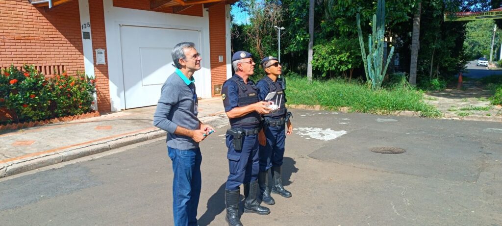 A imagem mostra três homens em uma rua asfaltada durante um dia ensolarado. Dois deles são agentes de segurança pública, provavelmente guardas municipais, vestidos com uniformes táticos azuis escuros, usando boinas e óculos escuros. Um dos agentes está segurando o controle remoto de um drone, enquanto ambos olham para o céu, provavelmente acompanhando o voo do equipamento. Ao lado deles, um homem vestido com uma camisa cinza de gola azul e calça jeans também observa o céu. A cena ocorre em uma área residencial, com casas de tijolos vermelhos e vegetação ao redor, incluindo um cacto alto e arbustos com flores vermelhas. Na parede de uma das casas, há uma placa com o número "135" e um aviso escrito "ENTULHO NESTE LOCAL", indicando que ali é permitido ou está proibido o descarte de entulho, conforme o contexto local. Ao fundo, uma rua se estende até uma área com árvores e um carro branco estacionado. A iluminação é forte, típica de um dia claro, com sombras definidas no chão.