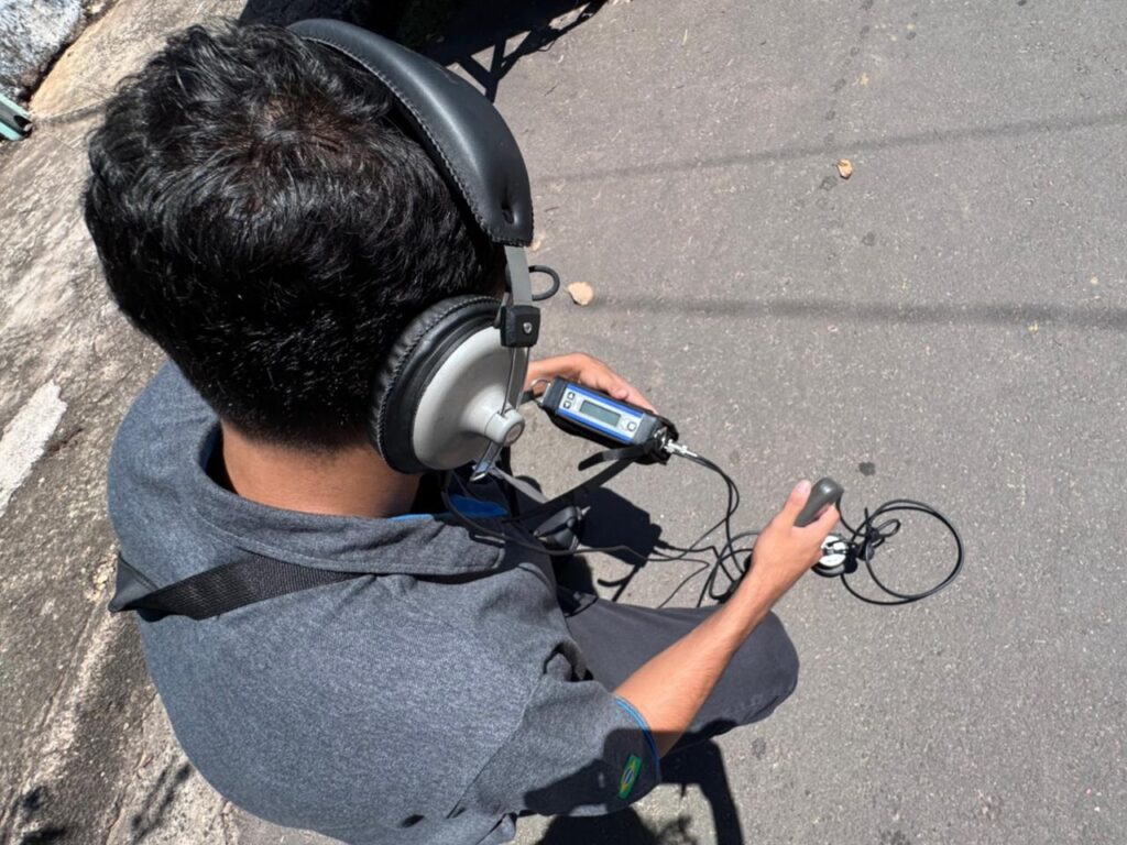 A imagem mostra uma pessoa vista de cima e por trás, agachada em uma superfície de asfalto. A pessoa usa uma camisa polo cinza com uma pequena bandeira do Brasil na manga esquerda e fones de ouvido grandes, cinza e pretos. Ela segura um dispositivo eletrônico com tela azul em uma das mãos e, com a outra, segura uma espécie de sensor ou aparelho conectado por fios ao dispositivo. A cena ocorre ao ar livre, sob luz solar intensa, que projeta uma sombra forte da pessoa no chão. O ambiente parece ser uma rua ou área pavimentada, e a atividade sugere que a pessoa está realizando algum tipo de medição ou inspeção técnica no solo.