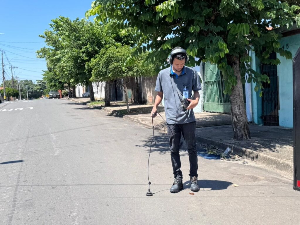 A imagem mostra um homem em uma rua asfaltada durante o dia, aparentemente realizando uma atividade de detecção de vazamentos ou inspeção de utilidades. Ele está usando uma camisa polo cinza com gola azul, calça escura e botas de trabalho pretas. O homem usa fones de ouvido grandes e segura um dispositivo eletrônico na mão direita, enquanto com a mão esquerda segura uma espécie de sensor ou bastão que encosta no chão. Ele também tem um crachá preso ao bolso da camisa. Ao fundo, há árvores verdes e um prédio azul claro, indicando que a cena ocorre em uma área residencial. A luz do sol está forte, projetando uma sombra nítida do homem no asfalto. A rua se estende à esquerda, onde um carro está estacionado. A imagem é bem focada e mostra detalhes do equipamento e da textura do pavimento e da vegetação.