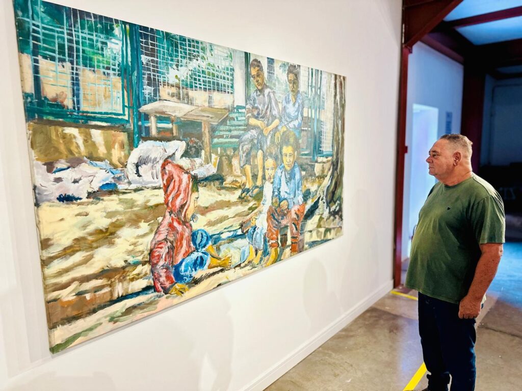 A imagem mostra um homem de meia-idade, vestido com uma camiseta verde, observando atentamente uma grande pintura a óleo exposta em uma parede branca de uma galeria de arte. A pintura retrata um grupo de crianças em um ambiente urbano ou de pobreza, com uma cerca de metal e destroços ao redor. As crianças estão sentadas, algumas no chão e outras em degraus ou bancos, e suas expressões são sérias ou neutras. A obra utiliza cores terrosas e pinceladas espessas, que conferem uma textura rugosa à pintura. O ambiente da galeria é claro e moderno, com piso de concreto polido e paredes brancas, e há uma porta aberta ao fundo que leva a um espaço mais escuro. A cena transmite uma atmosfera de reflexão sobre a infância em contextos difíceis.
