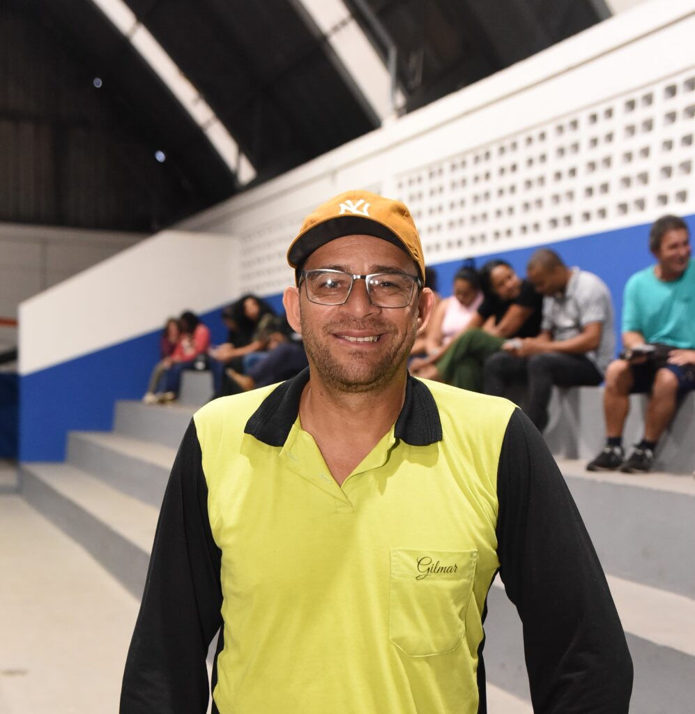 A imagem mostra um homem sorridente, de meia-idade, usando óculos retangulares e um boné laranja com a logo branca "NY". Ele veste uma camisa polo de manga longa, com o corpo em verde limão e as mangas e gola pretas. Na parte do peito da camisa, está bordado o nome "Gilmar". O homem está em um ambiente interno que parece ser uma quadra ou ginásio esportivo, com arquibancadas de concreto ao fundo onde algumas pessoas estão sentadas. A parede atrás das arquibancadas é branca com uma faixa azul e uma seção com um padrão decorativo. O teto é alto e escuro, com estrutura metálica aparente. A iluminação é clara, típica de locais esportivos cobertos. A atmosfera geral da foto é informal e amigável.