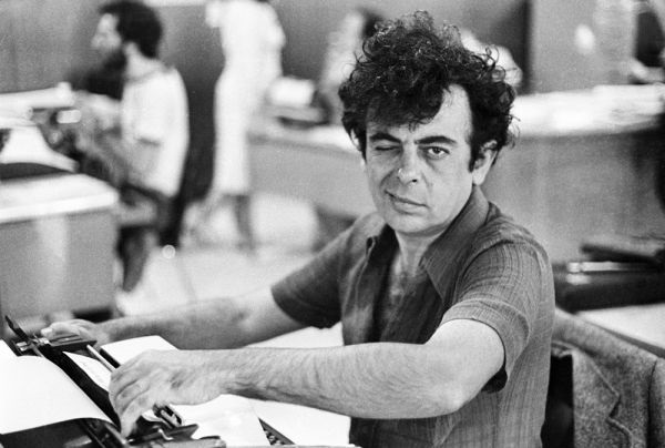 A imagem mostra um homem de meia-idade, com cabelo escuro e encaracolado, sentado em frente a uma máquina de escrever antiga, em um ambiente de escritório. Ele está sorrindo e piscando para a câmera, transmitindo uma expressão descontraída e amigável. O cenário ao redor é um escritório típico, possivelmente de uma redação ou ambiente administrativo, com móveis e outras pessoas desfocadas ao fundo. A fotografia é em preto e branco, com uma textura que lembra filmes antigos, sugerindo que a imagem foi tirada entre as décadas de 1960 e 1970. A iluminação é suave e natural, realçando os detalhes do rosto do homem e da máquina de escrever.