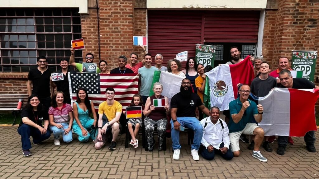 A imagem mostra um grupo grande de pessoas reunidas em frente a uma parede de tijolos vermelhos com uma grande porta de metal vermelha ao centro. O grupo está posicionado em duas filas principais: algumas pessoas estão agachadas na frente, enquanto a maioria está em pé atrás. Eles seguram várias bandeiras nacionais, incluindo uma grande bandeira dos Estados Unidos à esquerda, uma bandeira do México à direita, e uma bandeira da França mais à direita. Também há bandeiras menores de países como Espanha, França, Reino Unido, Alemanha e Itália, seguradas por diferentes pessoas. O grupo é diverso, composto por homens e mulheres de várias idades e etnias, todos com roupas casuais, como camisetas e jeans. Atrás deles, pendurados na parede, há dois banners verdes com o logo "CPP" e a frase "QUER PRATICAR IDIOMAS DE GRAÇA?". A atmosfera parece ser alegre e de celebração, provavelmente um encontro ou evento relacionado à prática de idiomas. O chão é pavimentado com tijolos intertravados, e uma janela com grade metálica aparece na lateral esquerda da imagem.