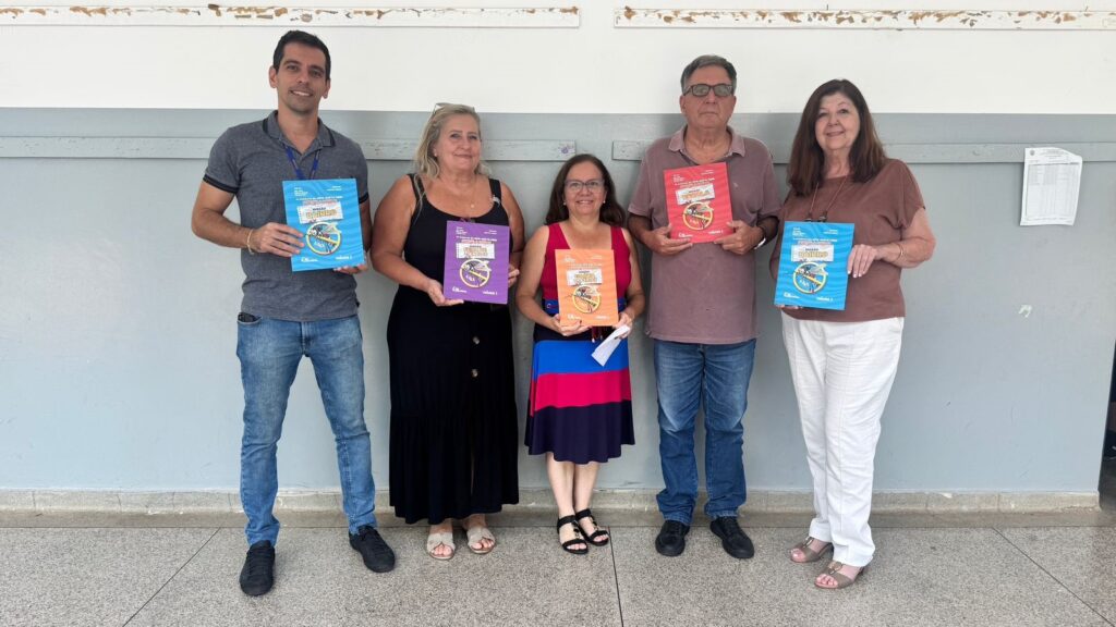 A imagem mostra cinco adultos alinhados, sorrindo para a câmera, em um ambiente interno com parede clara ao fundo. Cada um segura um livro colorido, com capas em azul, roxo, laranja e vermelho, que parecem ser materiais educativos. Os livros exibem títulos como "MISSÃO BAIRRO", "MISSÃO FEIRA DE CIÊNCIAS", "MISSÃO COLÔNIA DE FÉRIAS" e "MISSÃO ESCOLA", indicando que fazem parte de uma série de volumes educacionais. As pessoas estão vestidas de forma casual, e o ambiente aparenta ser uma escola ou centro comunitário. A atmosfera é amigável e colaborativa, sugerindo um contexto de apresentação ou divulgação de projetos educativos relacionados à ciência ou saúde pública.