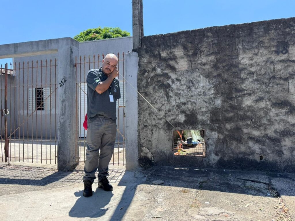 A imagem mostra um homem em pé, ao ar livre, próximo a uma parede de concreto desgastada e um portão metálico com barras verticais. O homem, calvo e com bigode, veste uma camisa polo cinza, calça cinza e botas pretas. Ele está segurando um fio fino próximo ao ouvido, como se estivesse escutando algo, enquanto a outra ponta do fio está conectada a uma pequena abertura quadrada na parede, onde é possível ver alguma tubulação ou equipamento. Ao fundo, através do portão, há um edifício com uma janela. A parede ao lado direito da imagem está marcada com o número "48". A cena ocorre em um dia ensolarado, com sombras nítidas no chão. A imagem transmite a ideia de que o homem está realizando algum tipo de inspeção ou manutenção.