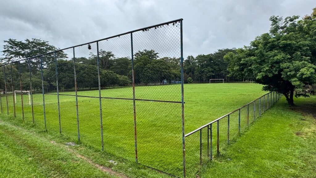 A imagem mostra um campo de futebol gramado cercado por uma cerca de arame galvanizado. O céu está nublado, com nuvens cinzentas que indicam um dia provavelmente chuvoso ou com pouca luz. Ao fundo, é possível ver árvores verdes que delimitam o espaço do campo. Há dois gols de futebol visíveis, um mais próximo à esquerda e outro ao fundo, indicando que o campo é usado para jogos formais. A cerca tem diferentes alturas em alguns trechos, e o ambiente geral transmite uma sensação de tranquilidade e simplicidade, típica de um campo ao ar livre em uma área pública ou escolar.