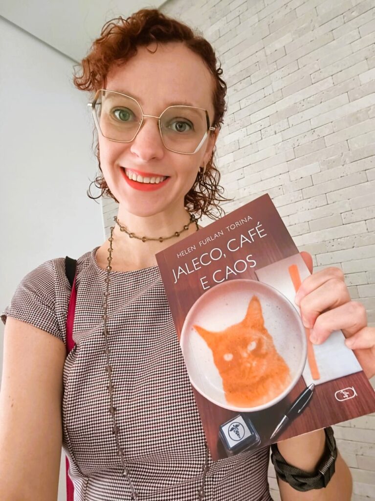 A imagem mostra uma mulher sorridente, de cabelos cacheados e ruivos, usando óculos de armação dourada em formato hexagonal. Ela veste uma blusa de padrão xadrez pequeno em tons de cinza e branco, e usa um colar de contas coloridas. A mulher está segurando um livro próximo ao rosto, intitulado "Jaleco, Café e Caos", escrito por Helen Furlan Torina. A capa do livro tem uma imagem de uma xícara de café vista de cima, com uma arte em forma de gato no leite, ao lado de uma caneta e um carimbo médico. O fundo da foto é composto por uma parede branca, metade texturizada em tijolos aparentes e a outra metade lisa. A iluminação é clara, destacando bem o rosto da mulher e o livro que ela segura. Ela também usa um bracelete escuro no pulso esquerdo. A expressão dela é alegre e confiante.