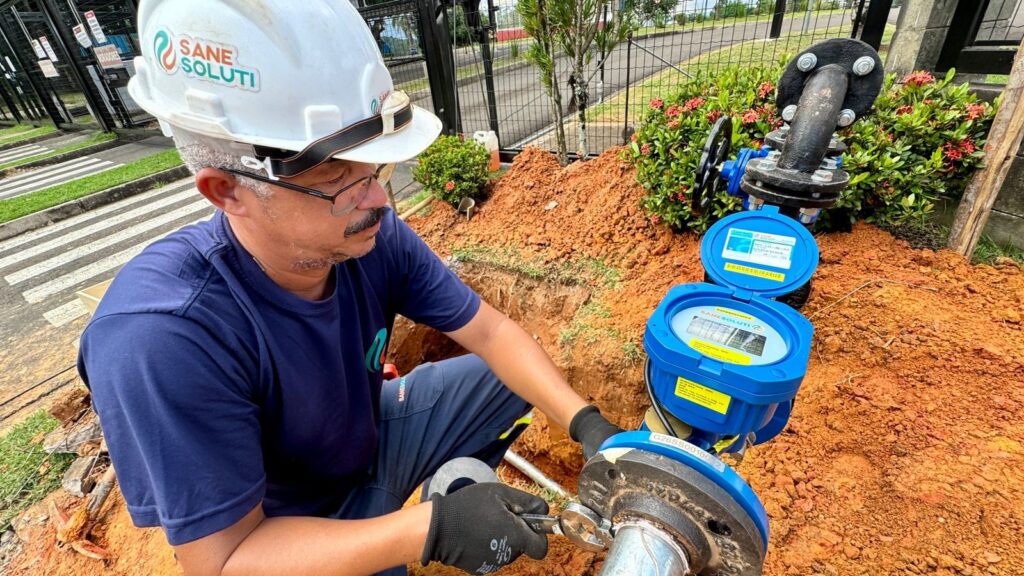 A imagem mostra um trabalhador masculino realizando a instalação ou manutenção de um sistema de medição de água em um ambiente externo. Ele está agachado ao lado de um pequeno buraco no chão, onde há um conjunto de tubos de metal pretos com grandes parafusos e uma caixa azul que parece ser um medidor de água digital, com a tampa aberta. O trabalhador usa um capacete branco com o logo "SANE SOLUTI", uma camiseta azul escura, calças de trabalho também azul escuro e luvas pretas de proteção. Ele está focado em apertar um parafuso na flange do tubo, utilizando uma ferramenta. Ao fundo, há uma cerca preta, plantas verdes com flores vermelhas e uma área gramada, sugerindo que o local é próximo a uma calçada ou entrada de uma residência ou estabelecimento. A cena transmite um ambiente de manutenção ou instalação de infraestrutura de saneamento básico, com destaque para o cuidado e profissionalismo do trabalhador.
