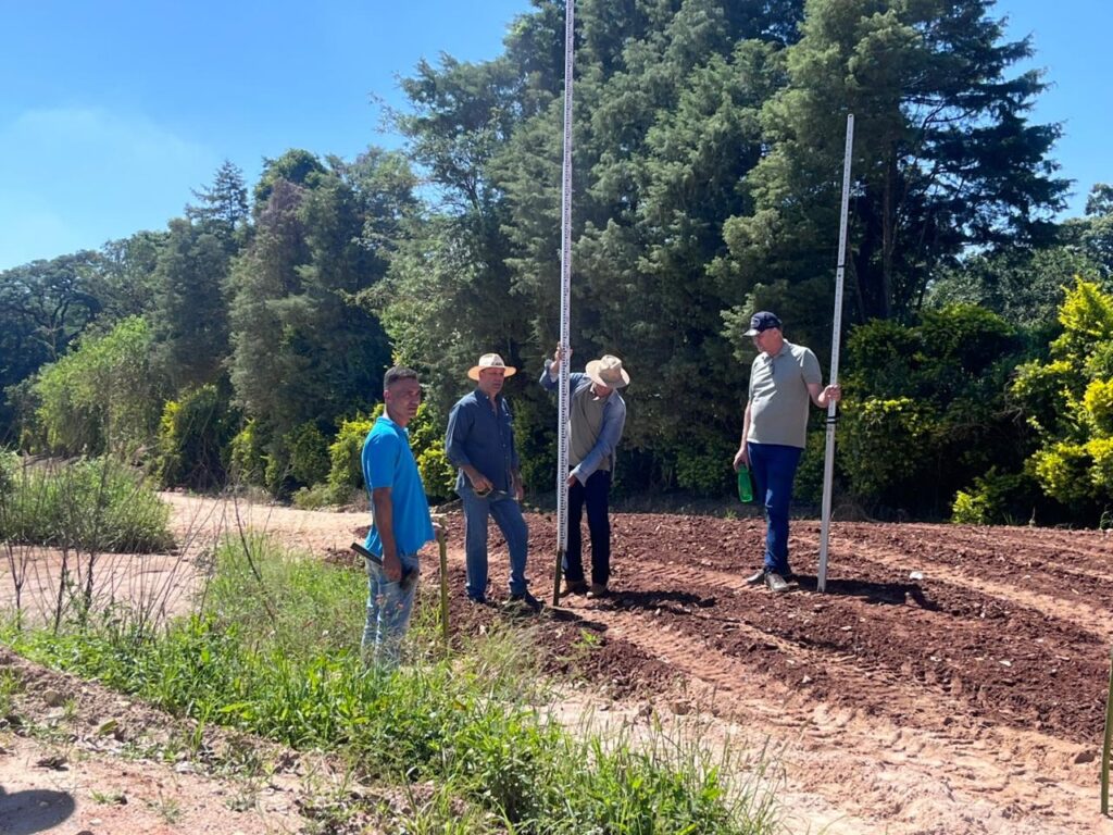 A imagem mostra quatro homens em um campo de terra arada, possivelmente em uma atividade de levantamento topográfico ou planejamento agrícola. O solo é de cor avermelhada e parece recentemente trabalhado, com marcas de pneus visíveis. Três dos homens estão posicionados no centro e à direita da imagem, segurando equipamentos de medição, como réguas topográficas longas, usadas para medir alturas ou distâncias. Um deles usa um chapéu e camisa azul, outro chapéu e camisa cinza, e o terceiro está com boné e camisa cinza clara. O quarto homem, à esquerda, veste uma camiseta azul e está de pé sobre uma área com grama, observando os outros. Ao fundo, há uma linha de árvores verdes sob um céu azul claro, indicando um ambiente rural em um dia ensolarado. A cena transmite uma atividade técnica ao ar livre, relacionada à agricultura ou construção.