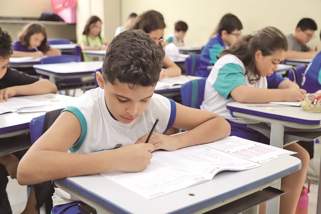 A imagem mostra uma sala de aula com várias crianças sentadas em carteiras brancas com detalhes azuis, concentradas em uma atividade escolar. Em destaque, um menino com cabelo cacheado escuro e pele clara está focado escrevendo em um caderno, usando uma camiseta branca com detalhes em azul e verde. Ao fundo, outras crianças também estão envolvidas na mesma atividade. A iluminação é natural e clara, e o ambiente transmite uma atmosfera de concentração e aprendizado. O caderno que o menino está usando possui a logo "Saeb", que indica que eles estão provavelmente realizando uma avaliação escolar padronizada no Brasil. A profundidade de campo da foto destaca o menino em primeiro plano, enquanto o restante da sala fica levemente desfocado.