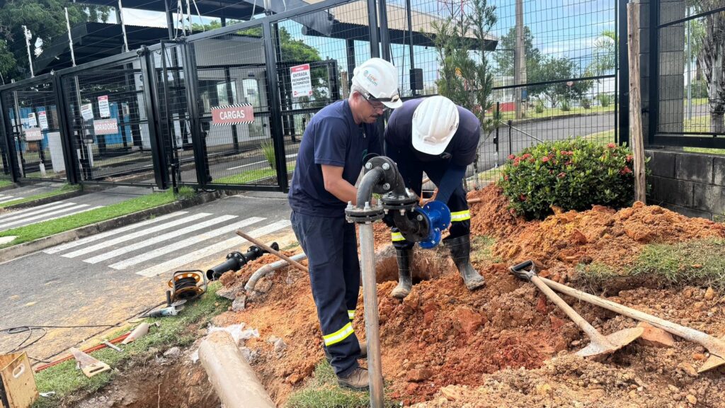 A imagem mostra dois trabalhadores realizando manutenção em um sistema de tubulação subterrânea ao lado de uma entrada com portão. Ambos estão usando capacetes brancos e uniformes azuis escuros com faixas refletivas amarelas nas pernas. Um dos trabalhadores está em pé, manipulando uma tubulação metálica vertical que sai do solo, enquanto o outro está agachado dentro de uma escavação, trabalhando em uma válvula azul conectada à tubulação preta. O chão ao redor da escavação está revolvido, com terra avermelhada acumulada e ferramentas como pás e picaretas espalhadas. Próximo à calçada, há um equipamento elétrico e cabos sobre a grama. Ao fundo, vê-se um portão metálico preto com placas indicativas em português: "VISITANTES", "MORADORES", "CARGAS" e um aviso de que a entrada possui fechamento automático, permitindo a passagem de um veículo por vez. Além do portão, há uma pequena guarita, uma rua pavimentada e vegetação. A cena está iluminada pela luz natural do dia, com foco na atividade técnica dos trabalhadores, mostrando o trabalho detalhado de manutenção das tubulações no local.
