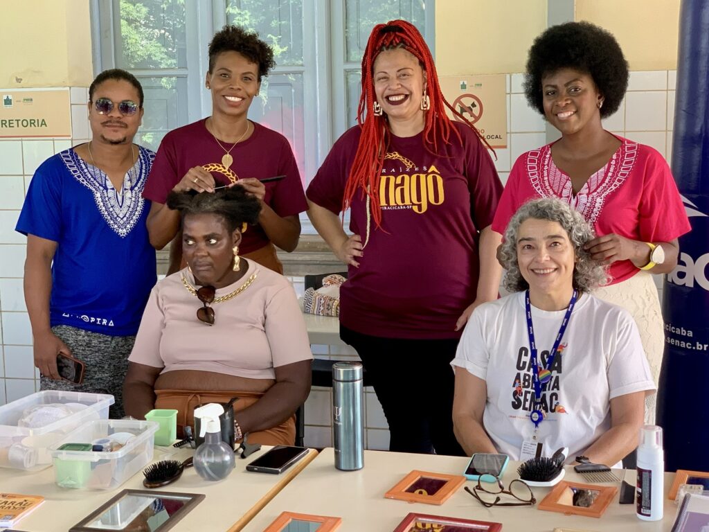 A imagem mostra um grupo de seis pessoas em um ambiente interno, provavelmente uma sala de aula ou oficina, com uma mesa à frente contendo diversos materiais de cabeleireiro, como escovas, pentes, espelhos e frascos de spray. Quatro mulheres estão em pé, enquanto duas estão sentadas. Uma das mulheres em pé está penteando o cabelo de uma das sentadas. As pessoas parecem estar participando de uma atividade prática relacionada a cuidados capilares ou estética. Na parede ao fundo, há uma placa com a palavra "REITORIA" e outras inscrições em português, indicando que o local é uma instituição educacional. Algumas das pessoas vestem camisetas com inscrições, como "RAIZES nagô" e "CASA ABERTA SENAC", sugerindo que o evento é promovido pelo SENAC em Piracicaba-SP. O ambiente é bem iluminado, com janelas ao fundo, e o clima parece ser de aprendizado e colaboração.