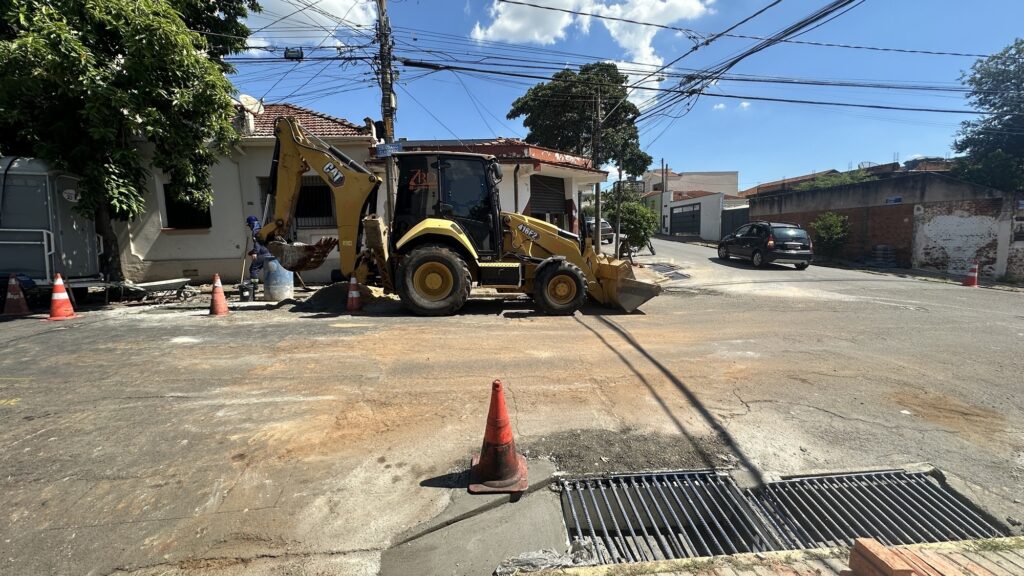 A imagem mostra uma máquina retroescavadeira amarela da marca Caterpillar (modelo 416F2) estacionada em uma esquina de rua residencial, aparentemente realizando trabalhos de manutenção ou reparo na via. Em volta da máquina, há cones de sinalização laranja que delimitam a área de trabalho, além de um barril azul e uma pequena pá de mão. Um trabalhador com roupa azul e capacete está próximo à retroescavadeira, indicando que o serviço está em andamento. A rua é asfaltada e há uma grande árvore verde ao lado esquerdo da imagem. Ao fundo, vê-se um carro preto se afastando. A placa da rua indica “R. CRISTIANO CLEOPATH”. O céu está claro com poucas nuvens, e há fiação elétrica visível sobre as construções baixas que cercam a área. A cena transmite um ambiente urbano tranquilo em um dia ensolarado.