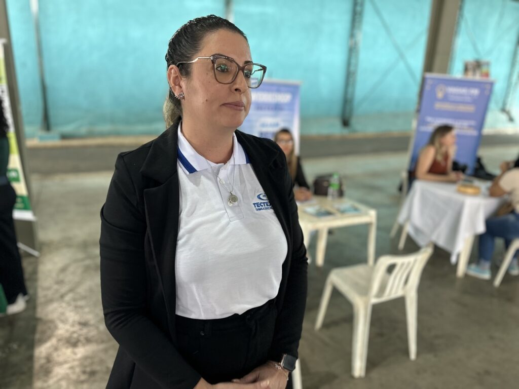 A imagem mostra uma mulher em primeiro plano, vestindo uma camisa polo branca com detalhes em azul e um blazer preto por cima. Ela usa óculos e um colar com um pingente redondo, além de um relógio inteligente no pulso. A expressão dela é séria e confiante. Ao fundo, desfocado, há várias pessoas sentadas em cadeiras brancas, em um ambiente que parece ser um evento corporativo ou feira de empregos, com banners azuis e mesas brancas. Na camisa da mulher, está estampado o nome "TECTEX EMBALAGENS", indicando que ela provavelmente representa essa empresa no evento. O ambiente é interno, com paredes de cor azul esverdeada e iluminação suave. A foto foca nitidamente na mulher, deixando o fundo desfocado para destacar a pessoa.