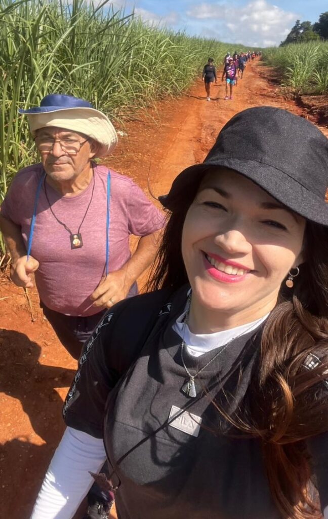 A imagem mostra uma mulher sorridente tirando uma selfie enquanto caminha por uma trilha de terra vermelha. Ela está usando um chapéu preto estilo bucket, uma camiseta preta da marca FILA com mangas brancas por baixo, e um colar com um pingente religioso. Ao lado dela, um homem mais velho, usando um chapéu de abas largas bege com detalhes azuis, óculos e uma camiseta rosa claro, também caminha pela trilha. Eles estão em um ambiente rural, cercados por canaviais altos que margeiam o caminho. Ao fundo, há um grupo de pessoas caminhando, sugerindo que fazem parte de uma caminhada coletiva ou peregrinação. O céu está claro com algumas nuvens brancas, e a luz do sol ilumina a cena, criando um ambiente alegre e tranquilo.
