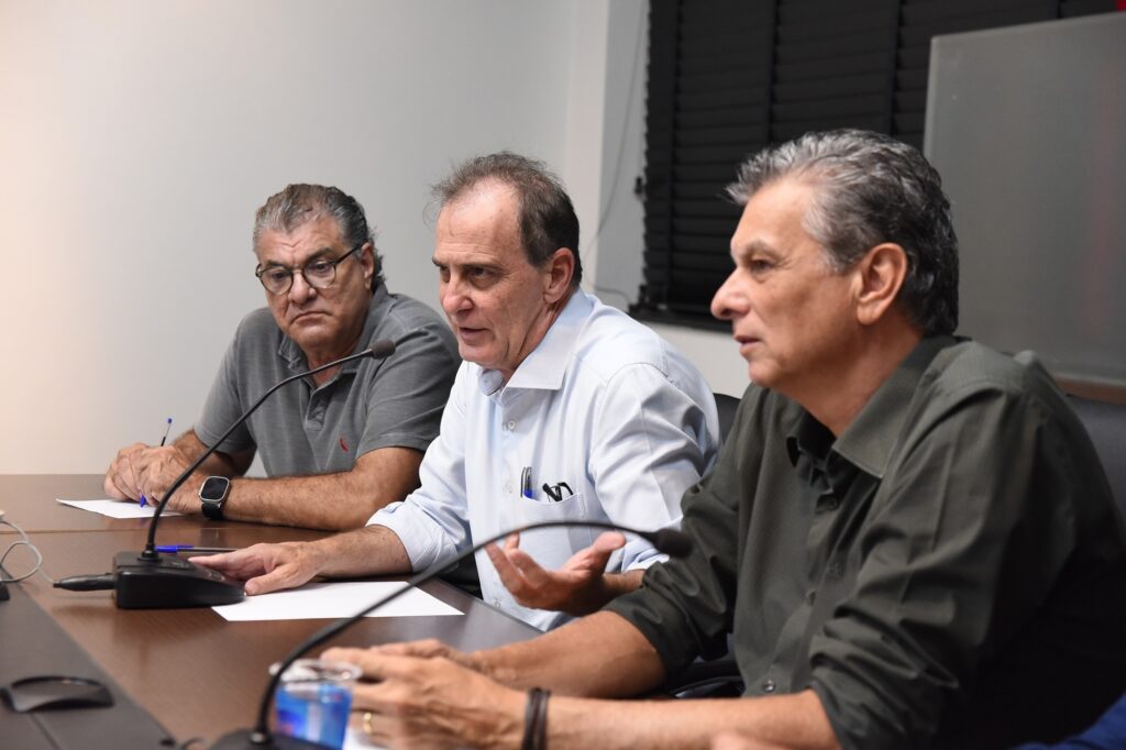 A imagem mostra três homens sentados em uma mesa de reunião, em um ambiente interno, provavelmente um escritório ou sala de conferências. Eles estão posicionados lado a lado, vistos em um ângulo diagonal. O homem no centro, que parece estar falando, veste uma camisa azul clara e segura uma caneta, demonstrando estar envolvido na conversa. O homem à esquerda, com uma camisa polo cinza e óculos, observa atentamente enquanto segura uma caneta azul. O homem à direita, com uma camisa verde escura, está de perfil, também atento à discussão. Sobre a mesa de madeira escura há papéis, canetas, microfones de mesa e um copo plástico com água. O ambiente é iluminado e tem uma parede branca ao fundo, com uma persiana preta à direita. A cena transmite uma atmosfera de reunião ou debate formal.
