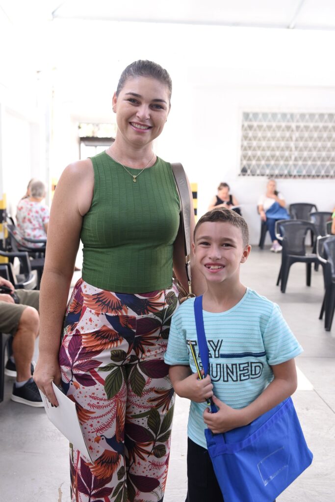 A imagem mostra uma mulher e um menino juntos, ambos sorrindo para a câmera. A mulher tem cabelo preso em um coque e veste uma blusa regata verde oliva e uma calça larga com estampa tropical colorida, com folhas e pássaros em tons de laranja, roxo e verde. Ela segura um documento branco na mão direita e carrega uma bolsa marrom no ombro. O menino veste uma camiseta listrada azul clara com a frase "STAY TUNED" estampada e carrega uma bolsa azul. Ele segura um lápis amarelo na mão. Eles estão em um ambiente bem iluminado, que parece ser uma área de espera, com pessoas sentadas ao fundo em cadeiras pretas. A atmosfera da foto é casual e alegre.